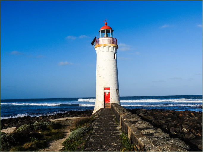 "Port Fairy lighthouse standing tall on the Shipwreck Coast, captured by photographer Barmah John. The lighthouse is bathed in warm light, with rugged coastal scenery in the background."